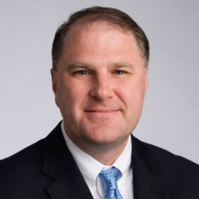 A headshot of a man in a black suit jacket against a light background.